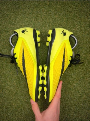 Gula Nike Hypervenom fotbollsskor med svart swoosh - Snygga gula fotbollsskor från Nike med svart swoosh och svart snörning. Skorna har en texturerad ovansida för bättre bollkontroll och är perfekta för dig som vill sticka ut på planen. Lätta och smidiga med platt sula och klassisk design. Hypervenom