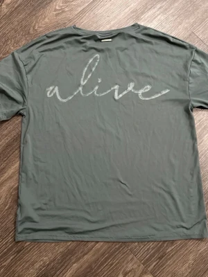 Sällsynt Gymshark x Whitney Simmons alive t-shirt - Snygg mörkgrön/grå t-shirt med rund hals och korta ärmar. På ryggen står det 'alive' i ett stort, handskrivet typsnitt. Tillverkad i mjuk bomull som känns skön mot huden. Perfekt för dig som gillar enkel men uttrycksfull stil.