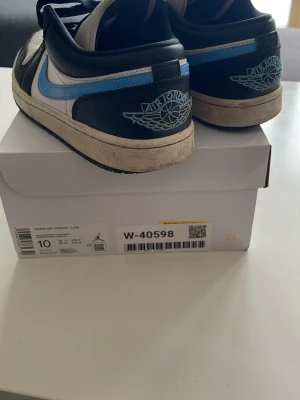 Nike Air Jordan 1 Low svart/blå/vit - Säljer ett par Nike Air Jordan 1 Low sneakers i svart, blått och vitt. Skorna har klassisk snörning, rund tå och den ikoniska Air Jordan-loggan broderad på hälen. 