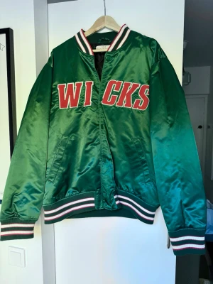 Grön bomber/baseballjacka med WICKS-tryck - Snygg grön bomberjacka från Urban Classics med stort rött och vitt WICKS-tryck på bröstet. Jackan har ribbade muddar med ränder i vitt, rött och grönt samt klassisk bomberkrage. Tillverkad i glansig polyester och har lång ärm.
