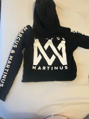 Svart Marcus & Martinus hoodie - Cool svart hoodie med stor vit Marcus & Martinus-logga på bröstet och text längs båda ärmarna. Croppad modell med huva och långa ärmar. Perfekt för dig som gillar streetstyle och vill sticka ut med en snygg merchtröja.