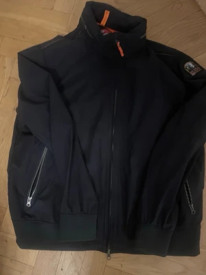 Svart vindjacka från Parajumpers - Snygg svart vindjacka från Parajumpers i softshell-material. Jackan har hög krage, dragkedja framtill och två sidofickor med dragkedja. Orangea detaljer och patch på ärmen ger en cool touch. Perfekt för blåsiga dagar.liten i storlek passar L