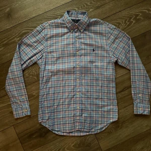 Rutig skjorta från Ralph Lauren - Klassisk rutig skjorta från Ralph Lauren i blått, vitt och rött. Skjortan har lång ärm, knappar framtill och button-down krage. Tillverkad i mjuk bomull och har den ikoniska broderade loggan på bröstet.