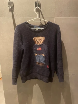 Mörkblå stickad tröja Polo Bear - Mörkblå stickad tröja från Polo Ralph Lauren med ikonisk björn på bröstet. Tröjan har rund halsringning, långa ärmar och ribbade muddar. Björnen bär jeans och tröja med amerikansk flagga. Perfekt för dig som gillar klassisk preppy-stil. Självklart äkta😀