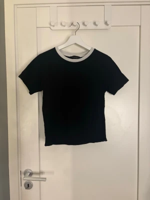 Svart t-shirt med vit krage - En stilren svart t-shirt med kontrasterande vit krage. Modellen är kortärmad och har en klassisk passform som funkar till det mesta. Perfekt basplagg för dig som gillar enkel och clean stil.