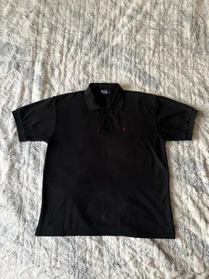 Svart pikétröja från Ralph Lauren - Klassisk svart pikétröja från Ralph Lauren med korta ärmar och krage. Storlek L. Tröjan har en liten röd broderad logga på bröstet och är tillverkad i bomull. Perfekt för en stilren och avslappnad look. T-shirten är i mycket gott skick och är använt ett fåtal gånger. Nypris är 1599kr.
