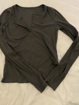 Svart långärmad topp från Dazy - En svart långärmad topp från Dazy med knappar framtill och en lätt v-ringad hals. Toppen har en enkel och clean design som passar till det mesta. Materialet känns mjukt och stretchigt, perfekt för en chill vardagslook.