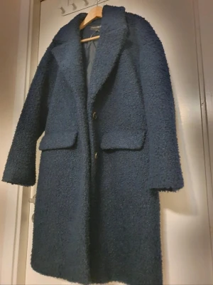 Mörkblå teddy kappa med fickor - Mysig mörkblå teddy kappa med stora slag och två "fejk-" framfickor. Kappan har en rak passform och stängs med knappar framtill. Perfekt för kyliga dagar och ger en trendig look med sin fluffiga textur.