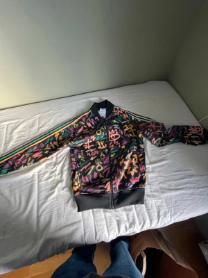 Adidas trackerjacka limitet graffitimönster - Säljer en Adidas bomberjacka i svart med färgglatt grafiskt mönster i neonfärger. Jackan har dragkedja, ribbade muddar och klassiska Adidas-ränder på ärmarna. Materialet är polyester och jackan har en normal passform. Perfekt för dig som vill sticka ut.