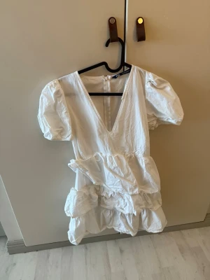 Vit studentklänning med puffärm - Söt vit kortklänning med djup v-ringning, puffärmar och flera volangdetaljer. Klänningen har en omlottkänsla och är tillverkad i ett lätt bomullstyg som passar perfekt för varmare dagar. Stängs med dragkedja i ryggen.