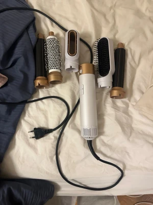 Multistyler hårverktyg med tillbehör - Säljer en multistyler för hår med fem olika utbytbara tillbehör. Verktyget är vitt och guldigt med svart sladd och passar för att skapa olika frisyrer. Inkluderar rundborste, fönmunstycke, två locktillbehör och en borste.