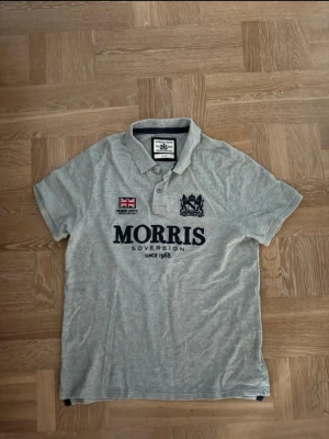  piké från Morris - Klassisk grå pikétröja från Morris  med broderad logga och brittisk flagga på bröstet. Tröjan har krage, knappar framtill och korta ärmar. Perfekt för en avslappnad men stilren look.