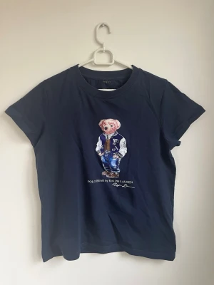 Mörkblå Polo Bear t-shirt Ralph Lauren - Snygg mörkblå t-shirt från Ralph Lauren med det ikoniska Polo Bear-motivet på bröstet. Klassisk passform med rund hals och korta ärmar. Tillverkad i mjuk bomull som känns skön mot huden. Perfekt för dig som gillar stilrena och lekfulla plagg.