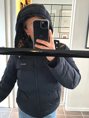 Vinterjacka från gant - Snygg mörkblå dunjacka från Gant med huva och dragkedja framtill. Jackan har quiltad design, två fickor och diskret logga på bröstet. Perfekt för kalla dagar när du vill hålla dig varm och stilren.