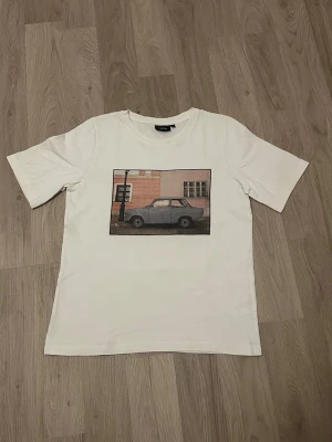 Vit t-shirt med biltryck från KappAhl - Vit t-shirt från KappAhl med ett coolt tryck av en bil framför en byggnad på bröstet. Klassisk rund hals och korta ärmar. Tillverkad i mjuk bomull som känns skön mot huden. Perfekt för dig som gillar streetstyle och unika prints.