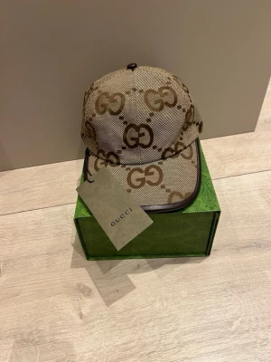 Beige keps med GG-mönster från Gucci - Snygg beige keps från Gucci med klassiskt GG-monogram i brunt över hela kepsen. Justerbar rem baktill i brunt läder och detaljer i guld. Tillverkad i canvasmaterial med exklusiv känsla. Perfekt accessoar för att lyfta din stil.