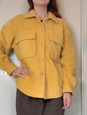 Gul Overshirt Jacka Fickor Tryckknappar Gina Tricot - Overshirt/jacka i en härlig gul färg med två stora bröstfickor och tryckknappar framtill. Jackan har en avslappnad passform, klassisk krage och är tillverkad i ett mjukt, fleece-liknande material. Perfekt för lager-på-lager, ensam som jacka och en cool vardagslook.