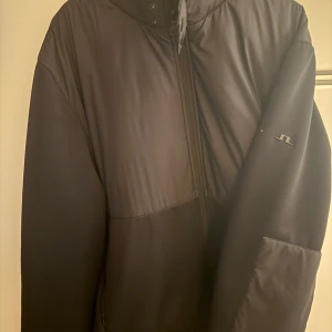 Svart vindjacka från J.Lindeberg - Snygg svart vindjacka från J.Lindeberg med diskret logga på ärmen. Jackan har hög krage, dragkedja framtill och elastiska muddar. Perfekt för dig som vill ha en stilren och sportig look.