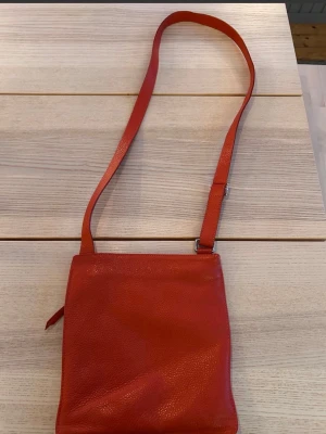 Röd cross body-väska - Snygg röd axelväska i skinn med justerbart axelband och dragkedja baktill. Väskan har en enkel, stilren design och passar perfekt för dig som vill ha en praktisk och trendig accessoar till vardagen. Väskan har ett fack med dragkedja på insidan samt ett fack med dragkedja på ena yttersidan.  Mått: 24x25 cm.  Väskan är i mycket gott skick och ser knappt använd ut.