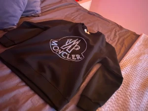 Svart Moncler sweatshirt med stor logga - Svart sweatshirt från Moncler i storlek S med en fet vit logga tryckt på bröstet. Tröjan har rund hals, långa ärmar och ribbade muddar. Perfekt för dig som gillar stilrena och exklusiva märken. Materialet känns mjukt och skönt mot huden.