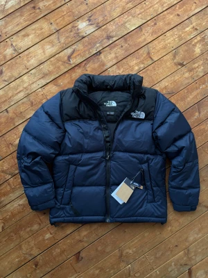 The North Face 1996 Retro Nuptse  - Blå/svart The North Face 1996 Retro Nuptse dunjacka i storlek S. Klassisk och varm vinterjacka med ikonisk boxig passform. Jackan är helt ny med prislapp kvar – perfekt för kalla dagar och tidlös i stilen.