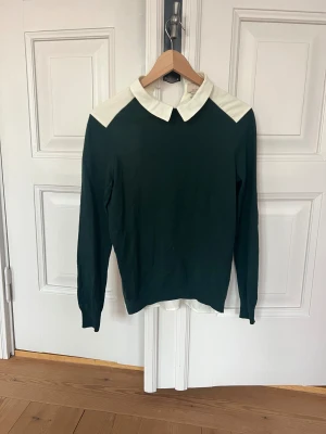 Grön tröja med vit krage från H&M - Stilfull grön långärmad tröja från H&M. Baksidan är i ett vitt tunnare tyg med panel och söt knapp vid nacken. Tröjan är i mjukaste ullblandning och har en klassisk välskräddad stil. Perfekt både vardag och mer uppklätt, supersnygg både till jeans, kostymbyxa och kjol, passar året om. Lätt att bära även under en kavaj. Modellen på bilderna är strl 36/S.