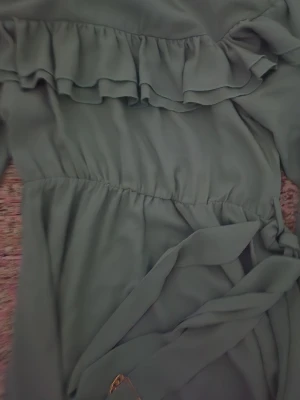 Ljusgrön långklänning med volang - Snygg ljusgrön långklänning med volangdetaljer över bröstet och långa ärmar med resår vid handlederna. Klänningen har ett knytband i midjan och är tillverkad i ett mjukt, flowy polyestertyg som ger en skön känsla. Perfekt för dig som gillar romantisk stil och vill sticka ut.