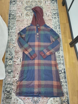 Rutig långklänning med huva - Långklänning med stor huva och knytband. Klänningen har ett rutigt mönster i blått, grönt, vinrött och beige, samt knappar framtill. Materialet känns som mjuk bomull.