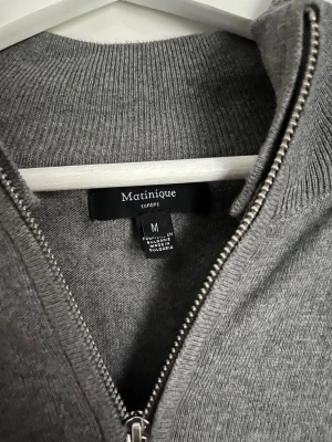 Grå kofta med dragkedja från Matinique - Snygg grå kofta från Matinique i storlek M. Modellen har hel dragkedja framtill, ribbad krage och muddar vid ärmslut och nederkant. Tillverkad i mjuk bomull, perfekt för lager-på-lager under höst och vår. Enkel och stilren design som funkar till många outfits.