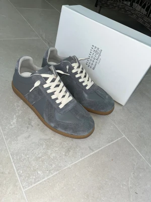 Maison Margiela grå sneakers strl 42 - Maison Margiela sneakers i grått med beige snörning och gummisula. Ovandelen är en mix av mocka och skinn, med klassisk rund tå och platt sula. Snygg kontrast mellan de olika grå nyanserna och den ljusa sulan. Perfekt för dig som gillar stilrena och exklusiva sneakers.