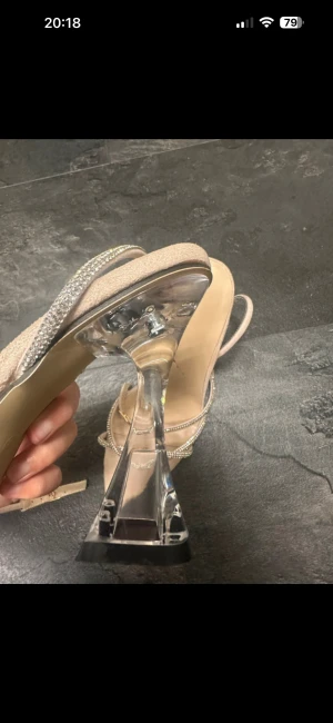 Beige sandaler med strass och klar klack - Säljer ett par snygga beige sandaler med spetsig tå och tunna remmar dekorerade med glittrande strass. Klacken är hög och i transparent plast, vilket ger en cool och modern vibe. Perfekt för dig som vill sticka ut med detaljer och stil.