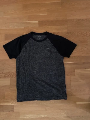Mörkgrå t-shirt från Hollister - Snygg mörkgrå t-shirt från Hollister med svarta raglanärmar och rund halsringning. T-shirten har en liten broderad Hollister-logga på bröstet och är tillverkad i mjuk bomullsmix. Perfekt för en avslappnad stil.