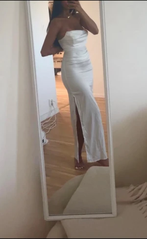 Vit axelbandslös långklänning med slits - Säljer en elegant vit långklänning med axelbandslös design och figurnära passform. Klänningen har en snygg slits framtill som ger en extra touch och är tillverkad i ett glansigt, satinliknande material. Perfekt för dig som vill sticka ut med en stilren look.