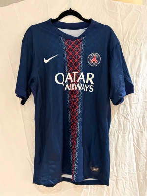 PSG João Neves  #87 - Paris Saint-Germain fotbollströja med João Neves och nummer 87 på ryggen. Tröjan är marinblå med geometriskt mönster i blått, vitt och rött, PSG-emblem på bröstet och Nike-logga. Tillverkad i lätt, ventilerande syntetmaterial. Snygg design för dig som älskar fotboll och PSG.