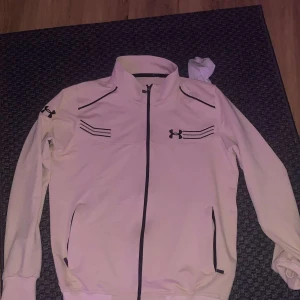 Vit vindjacka från Under Armour - Snygg vit/beige vindjacka från Under Armour med svarta detaljer och dragkedja framtill. Jackan har hög krage, svarta loggor på bröstet och ärmarna samt praktiska fickor med dragkedja. Perfekt för dig som gillar sportig och clean stil. Kommer med en svart under armour fixa så de blir en full tracksuit, helt ny har tags dock dem är borttagna.  Med byxorna kostar de 450kr utan 250kr