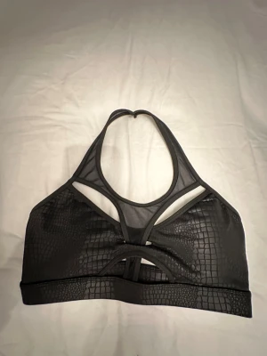 Svart sport bh med meshdetaljer - Säljer en svart sport bh med coolt krokodilpräglat mönster och meshdetaljer framtill och upptill. Toppen har en snygg cut-out design och bred kant nedtill. Perfekt för att lyfta din outfit med edgy vibes.