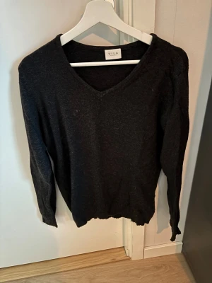 Mörkgrå stickad tröja från Vila - Snygg mörkgrå stickad tröja från Vila med v-ringad hals och långa ärmar. Tröjan har en klassisk, enkel design och är mjuk och skön mot huden. Perfekt för dig som gillar stilrena plagg med avslappnad passform.