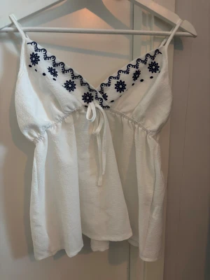 Vit linnetopp med blå broderi - Supersöt vit linnetopp med tunna axelband och blått broderat blommönster längs v-ringningen. Toppen har knyt framtill under bysten och är lätt utställd för en flowy känsla. Perfekt för varma dagar och har en fräsch, somrig vibe.