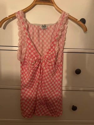 Rutig rosa linnetopp från D&G - Supersöt linnetopp från D&G med rosa och vit rutigt mönster, spetsdetaljer på axelbanden och en knytning framtill. Tunn och mjuk bomull, perfekt för varma dagar. D&G-logga tryckt längst ner. Snygg och girly vibe!