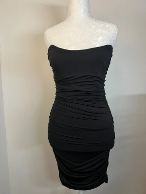 Klänning - Svart axelbandslös klänning med figurnära passform och rynkade detaljer längs hela klänningen samt dragkedja baktill. Storlek 36 (S) men passar även mig som vanligtvis bär XS. Använd en gång och i utmärkt skick 🖤