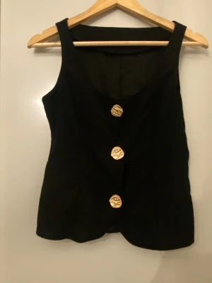 Svart och brun väst med guldknappar - Två stilrena västar i svart och brun med breda axelband och tre stora, strukturerade guldknappar framtill. Västarna har en rak passform och är tillverkade i ett mjukt bomullsmaterial. Perfekt för att ge din outfit en cool och elegant touch.