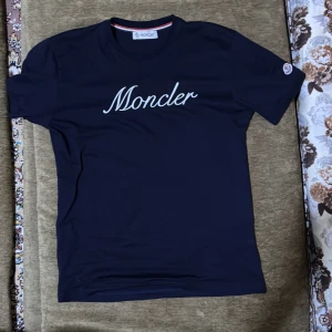 Mörkblå Moncler t-shirt  - Snygg mörkblå t-shirt från Moncler med vit broderad logga på bröstet och klassisk rund hals. T-shirten har korta ärmar och en liten Moncler-patch på ena ärmen. Tillverkad i mjuk bomull för en skön känsla.