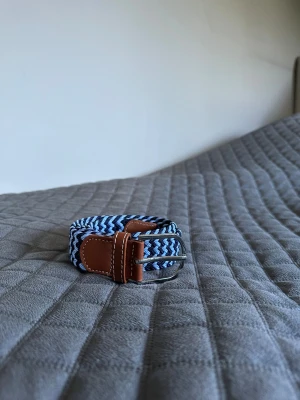 Blått och brunt flätat bälte - Säljer ett snyggt flätat bälte i marinblått och ljusblått med bruna skinn-detaljer och silvrigt spänne. Bältet har ett randigt mönster och är flexibelt tack vare den vävda designen. Perfekt för att ge outfiten lite extra stil.