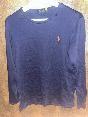Marinblå långärmad tröja Polo Ralph Lauren - Snygg marinblå långärmad tröja från Polo Ralph Lauren med klassisk rund hals och broderad logga på bröstet. Tröjan är i mjuk bomull och har en slim passform som sitter riktigt bra. Perfekt för dig som gillar stilrena och tidlösa plagg.