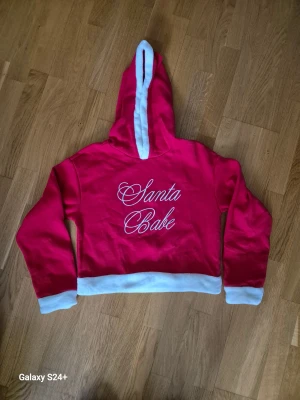 Röd croppad hoodie Santa Babe - Riktigt söt röd  hoodie med vit text 'Santa Babe'.Tröjan har huva och vita detaljer vid ärmslut, nederkant och huva. 