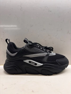 Dior b22 - Säljer ett par svarta Dior sneakers med chunky plattformssula och snygga silverdetaljer. Skorna har meshpaneler för extra ventilation, snörning och rund tå. Märket syns tydligt på sidan. Perfekt för dig som gillar exklusiv streetstyle.