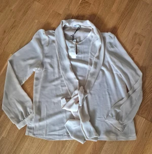 Mocha blus med knyt från Ida Sjöstedt - Säljer en snygg mocha-färgad blus från Ida Sjöstedt i silkeslen polyester. Blusen har långa ärmar, vacker knyt-detalj framtill och en mjuk, flowy passform. Perfekt för dig som gillar stilrena och feminina plagg med det lilla extra.