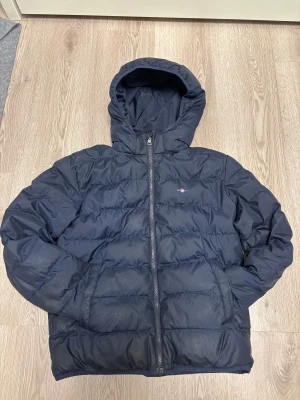 Mörkblå pufferjacka från GANT - Säljer en mörkblå pufferjacka från GANT med huva och dragkedja framtill. Jackan har quiltad design och två sidofickor. Perfekt för kalla dagar och har en clean look med diskret GANT-logga på bröstet. Passar dig som gillar stilren och funktionell ytterjacka. Jackan har dessutom extra ficka på insidan och är däremot inte så stor å puffig för de som gillar smalare jackor men fortfarande varma inför vintern. 
