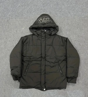 Svart pufferjacka med tryck på huvan - Svart pufferjacka med stor huva som har ett coolt tryck i grått. Jackan har dragkedja och knappar framtill, två snedställda fickor med dragkedja och ribbade muddar vid ärmsluten. Perfekt för kalla dagar och riktigt street.