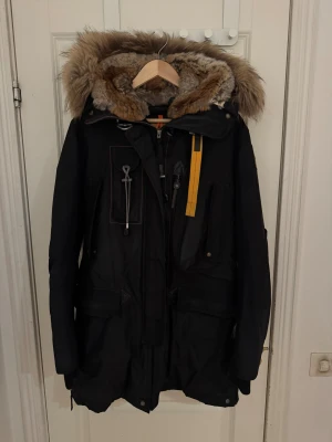 Svart Parajumpers parkajacka M - Parkajacka från Parajumpers i svart med stor fluffig päls runt huvan och coola detaljer som gul rem och snörning. Flera fickor, dragkedja och knappar framtill. Slim fit och riktigt varm för vintern. Perfekt för dig som vill ha en snygg och funktionell jacka.